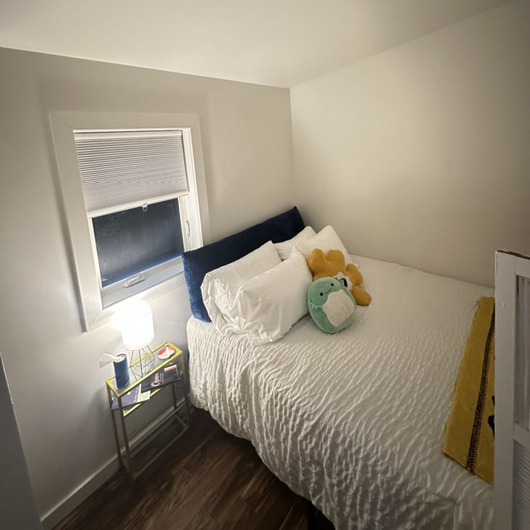 Sleeping area with bedside table, several pillows, colorful blanket, and screened window that opens. Not shown: spacious hinged-door closet. 