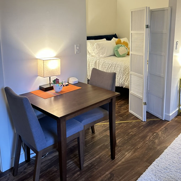 View of two seat dining area with lamp, with sleeping area in the distance.